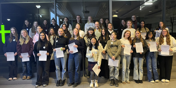 Eine große Gruppe von Mädchen und jungen Frauen steht in mehreren Reihen in einem Innenraum. Die meisten halten Papiere (Zertifikate) in den Händen. Die Kleidung ist vielfältig, darunter helle und dunkle Oberteile sowie Jeans. Im Hintergrund sind große Fenster und eine Decke mit sichtbaren Rohren und Lampen zu sehen.