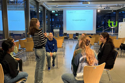 Six girls sit in a circle of chairs, a seventh girl stands in the centre. 