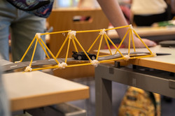 Modellbrücke aus Spaghetti mit kleinem Spielzeugauto auf einer gebogenen Fahrbahn zwischen zwei Tischen.