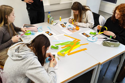 Four girls are sitting at a table with coloured cardboard strips in their hands. The workshop leader stands next to the table and watches what the girls are doing. 