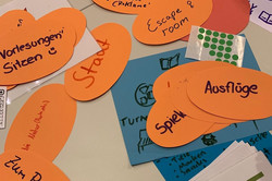 Mehrere orangefarbene, ovale Zettel mit handschriftlichen Notizen liegen auf einem Tisch, daneben eine Hand, die ein Dokument hält.