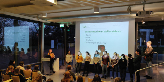 Students stand in front of a projector screen and introduce themselves to pupils.