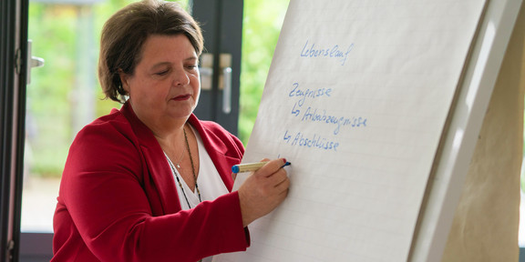 Eine Frau in rotem Sakko schreibt etwas an eine Flipchart