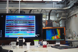 Zwei Bildschirme auf einem Tisch in einem Labor, einer zeigt ein farbiges Spektrum mit dem Text 'Multiwavelength Milky Way', der andere eine Wärmebildaufnahme mit Temperaturangaben, davor mehrere Becher und ein Mikrofon.