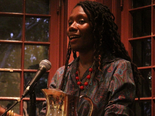 Person mit langen Zöpfen, die ein gemustertes Kleid und bunte Perlenarmbänder trägt, steht hinter einem Mikrofon mit einem Wasserkaraffe und einem Glas davor.