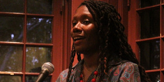 Person mit langen Zöpfen, die ein gemustertes Kleid und bunte Perlenarmbänder trägt, steht hinter einem Mikrofon mit einem Wasserkaraffe und einem Glas davor.