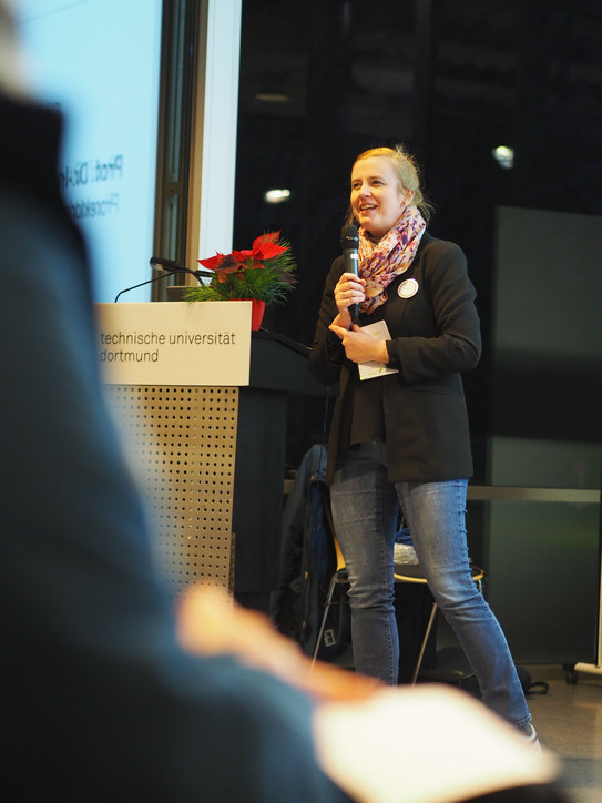 Petra Widerkehr steht neben einem Rednerpult. Sie hält ein Mikro in der Hand und spricht zu einem Publikum. 