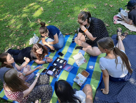 Auf einer Wiese liegt eine Picknickdecke. Sechs Mentees und Mentorinnen sitzen im Kreis und spielen das Kartenspiel "Tabu".