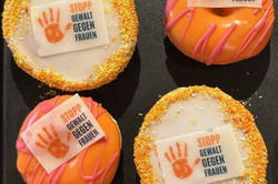 Photographed from above: two donuts with orange icing and two Americans with orange sprinkles on a black background. The pastries each have an edible sticker with the words ‘Stop violence against women’ on them