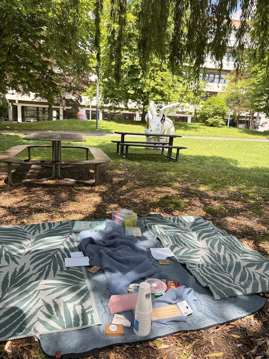 Picknickdecke mit Bechern, Snacks und Notizbüchern auf einer Wiese vor einem weißen Einhorn-Skulptur und Parkbänken unter Bäumen.