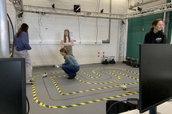 Auf dem Boden ist eine Strecke mit Tape abgeklebt. In der Mitte des Tapes fährt ein Lego-Roboter über eine schwarze Linie. Drei Mädchen steuern und beobachten den Roboter.