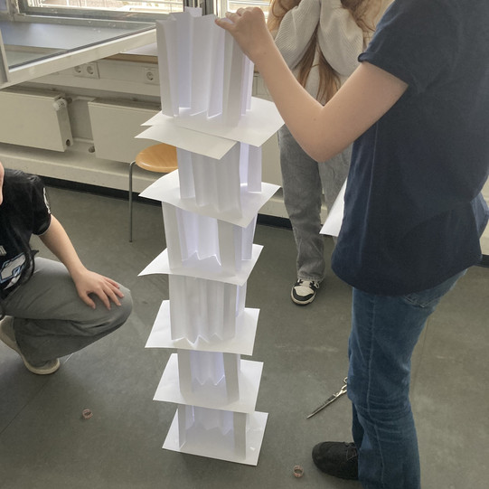 Mehrstöckiger Turm aus gefalteten und flachen weißen Papierbögen auf dem Boden, umgeben von drei Personen in einem Raum mit Fenster