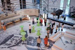 Gruppe von Menschen steht und sitzt an grünen Stehtischen und weißen Sofas in einer großen Halle mit technischen Geräten und Schautafeln.