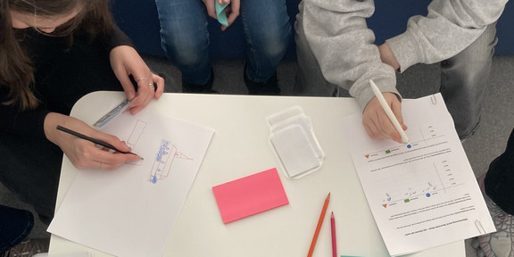 Von oben fotografiert: Zwei Personen sitzen an einem weißen Tisch und schreiben auf Papier mit Stiften, auf dem Tisch liegen bunte Notizzettel und ausgedruckte Blätter mit Diagrammen