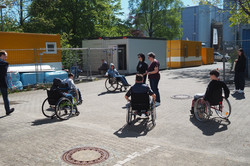 Mehrere Personen im Rollstuhl und zwei stehende Personen auf einem gepflasterten Hof mit Baucontainern im Hintergrund.