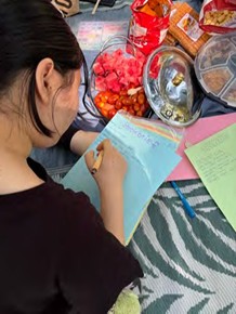 Person mit dunklen Haaren von hinten beim Schreiben auf einem hellblauen Blatt Papier auf einem gemusterten Untergrund mit mehreren bunten Gegenständen im Hintergrund.