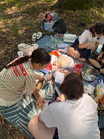 Gruppe von Personen sitzt auf einer Decke im Freien, umgeben von Taschen und Essen, unter einem Baum.