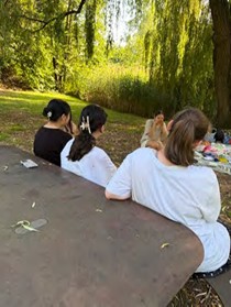 Drei Personen sitzen auf einer Parkbank mit Blick auf einen sonnigen, grünen Park mit Bäumen und Sträuchern.